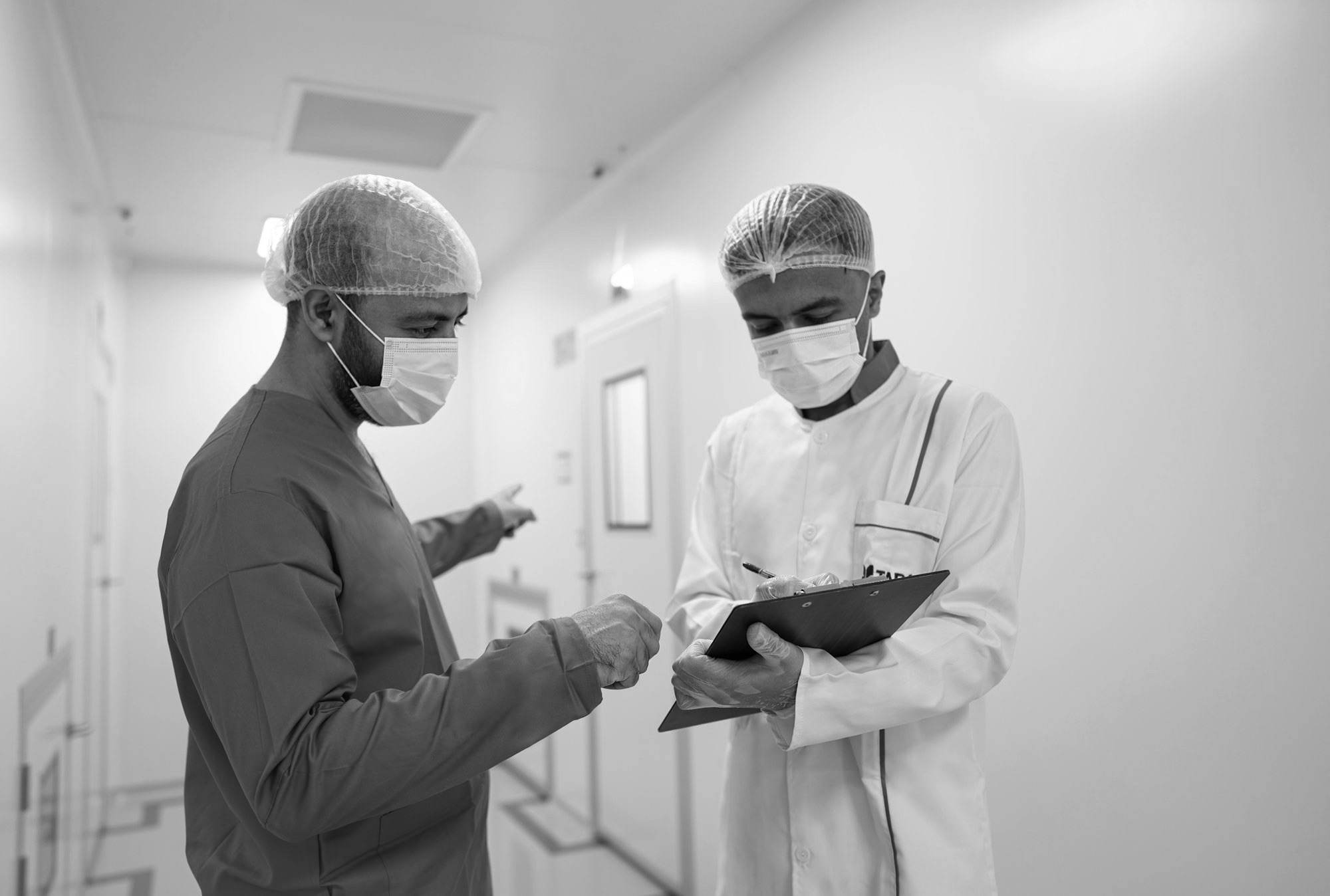 Two clinicians in masks and hair covers reviewing a clipboard in a hospital corridor